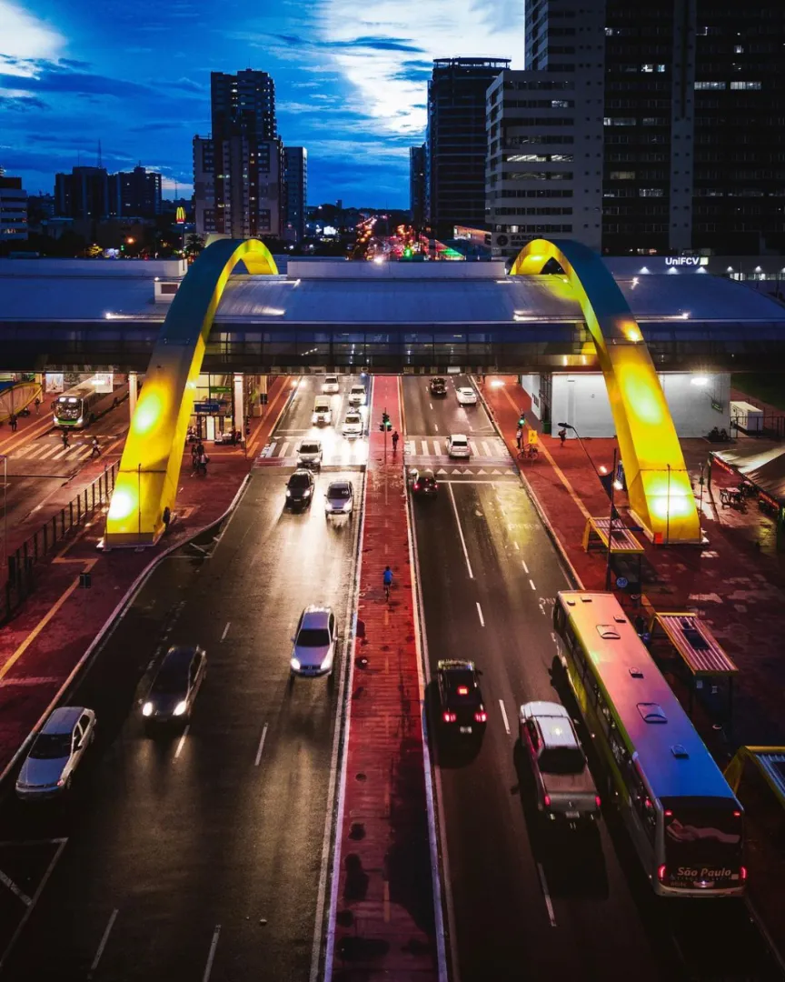 O entardecer agitado de Maringá. Carros, ônibus e pedestres passam por baixo do terminal urbano, formando uma mistura de cores e movimento.